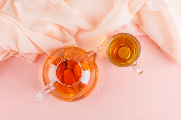 Thé à l'abricot en théière et tasse en verre sur table rose et textile, vue de dessus.