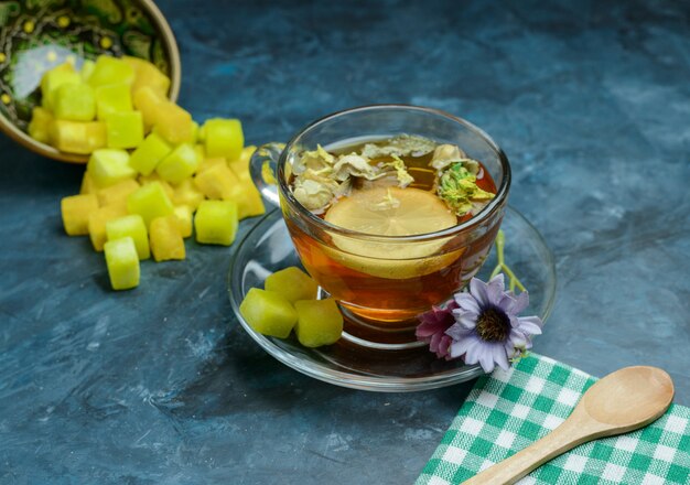 Thé aux herbes citronné avec des morceaux de sucre, une cuillère, un torchon dans une tasse sur une surface bleu foncé, vue en plongée.