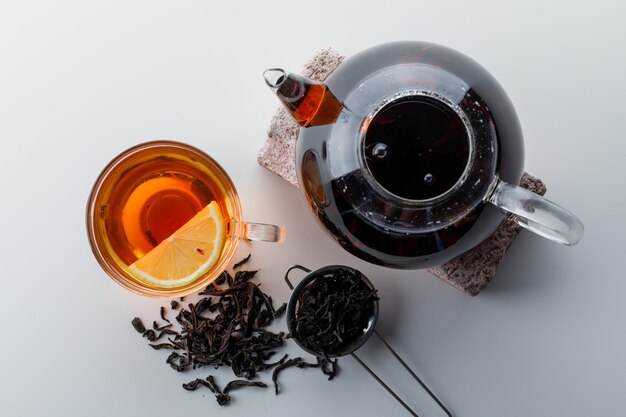 Thé citronné avec théière sur brique, passoire, thé sec dans une tasse sur une surface en dégradé blanc, vue de dessus