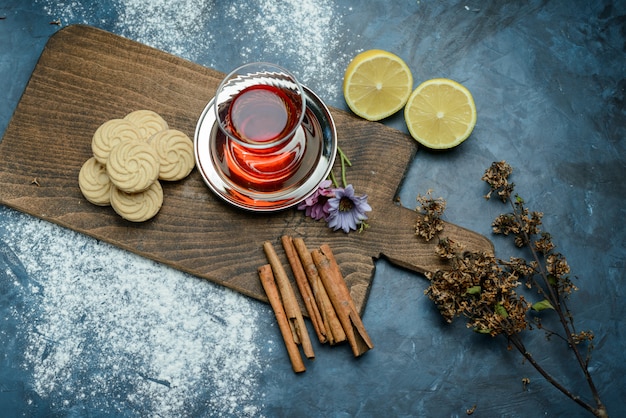 Thé dans un verre au citron, biscuits, herbes séchées, bâtons de cannelle, planche à découper vue de dessus sur la surface bleu grungy