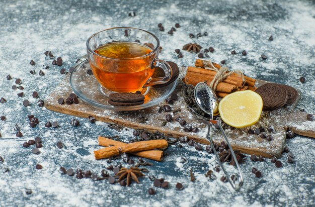 Thé avec farine, chips de choco, biscuits, épices, citron dans une tasse sur béton et planche à découper, vue grand angle.