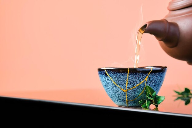 Le thé vert chaud est versé d'une théière en céramique dans un bol. Mise au point sélective sur la tasse bleue. La vapeur s'élèvera au-dessus de la tasse Tasse bleue en céramique récupérée, seconde vie des choses, recyclage ou kintsugi