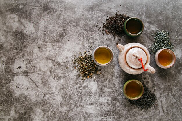 Théière en céramique entourée d'herbes séchées et de tasses à thé sur fond de béton