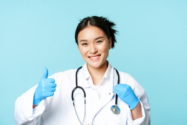 Thérapeute de femme asiatique souriante, médecin avec stéthoscope montre les pouces vers le haut, debout sur fond bleu.