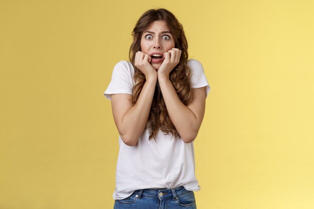 Timide insécurité intense haletant jeune femme séduisante effrayée soupirant choqué effrayé tenir la main face à se ronger les ongles terrifié exprimer la peur debout secoué fond jaune regarder la caméra.