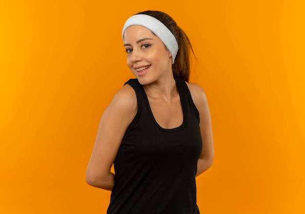 Timide jeune femme de remise en forme sportswear avec bandeau avec sourire sur le visage debout sur un mur orange