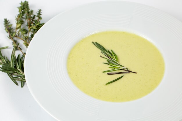 Tir d'une assiette creuse avec soupe à la crème de courgettes sur une table blanche décorée de plantes vertes