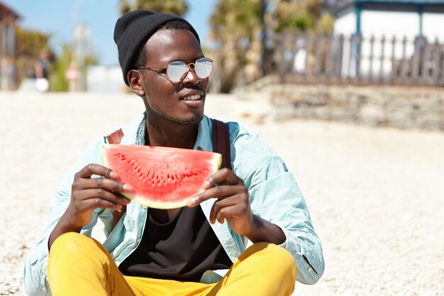 Tir d'été en plein air d'un bel homme à la peau sombre dans des vêtements à la mode et des lunettes de détente sur la plage pendant la journée, assis sur des galets avec une tranche de pastèque juteuse, profitant des fruits mûrs et du temps chaud