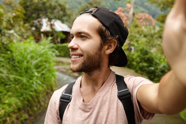 Photo gratuite tir extérieur d'un jeune hipster heureux portant un sac à dos et une casquette de baseball prenant un autoportrait, souriant et regardant ailleurs. beau voyageur marchant le long de la route de campagne