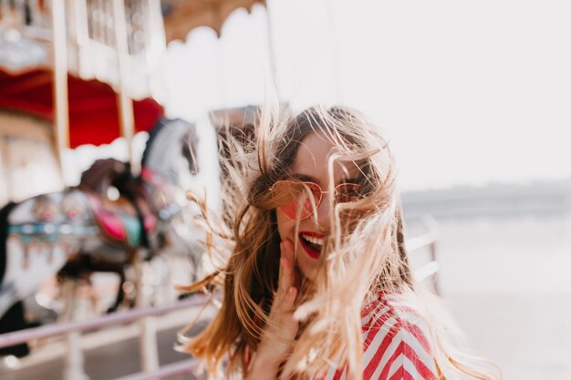 Tir extérieur d'une jolie fille heureuse exprimant des émotions positives. Jeune femme rêveuse à lunettes de soleil posant avec plaisir dans un parc d'attractions.