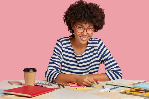 Tir horizontal d'agréable multiplicateur avec coupe de cheveux afro, utilise un crayon, bénéficie d'une boisson chaude