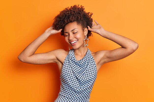 Tir horizontal d'une femme afro-américaine optimiste garde la main sur les cheveux bouclés ferme les yeux avec satisfaction sourit largement