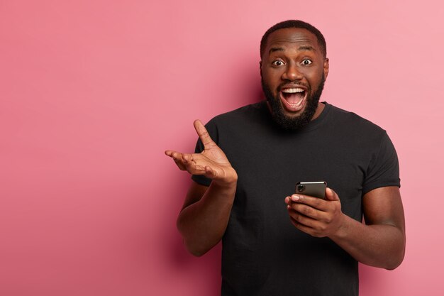 Tir horizontal d'heureux homme noir utilise un téléphone mobile moderne, des gestes avec la main, s'exclame d'émotions positives, obtient un beau message, porte un t-shirt noir