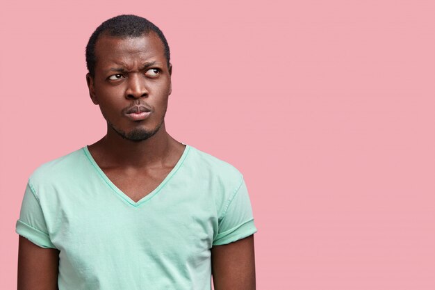 Tir horizontal de jeune homme afro-américain attrayant réfléchi à la peau foncée, habillé en t-shirt décontracté, pose contre rose