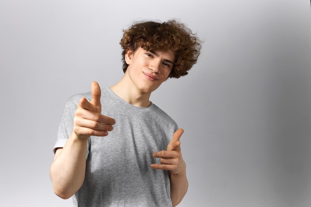 Tir horizontal de mignon jeune homme confiant avec un visage lisse et une coiffure frisée portant des vêtements décontractés posant sur fond gris pointant les doigts avant à la caméra, souriant, être fier de vous