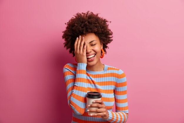 Tir à l'intérieur d'une belle femme aux cheveux bouclés fait visage plam, ferme les yeux, rit positivement, sourit largement, a une conversation heureuse avec un ami, apprécie la boisson à la caféine le matin, habillé avec désinvolture