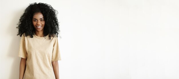 Tir intérieur de la belle jeune femme africaine avec une coiffure afro vêtue d'un t-shirt oversize décontracté souriant joyeusement debout au mur blanc blanc