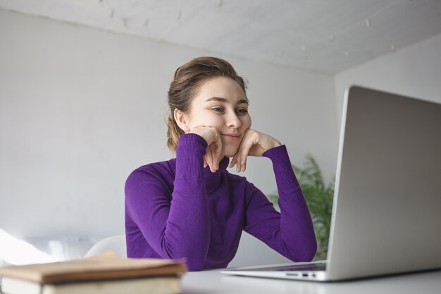 Tir intérieur de la belle jeune femme en haut à manches longues violet assis au bureau devant un ordinateur portable ouvert, regardant le blog vidéo