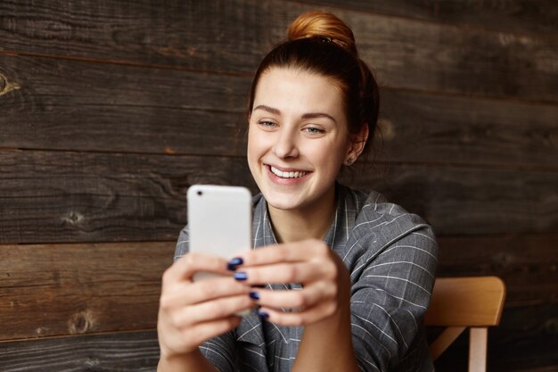 Tir intérieur de la belle jeune femme souriant joyeusement tout en envoyant des SMS à des amis