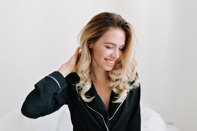 Photo gratuite tir intérieur de belle jeune mannequin aux cheveux blonds habillés de vêtements de nuit assis dans le lit avec un sourire merveilleux