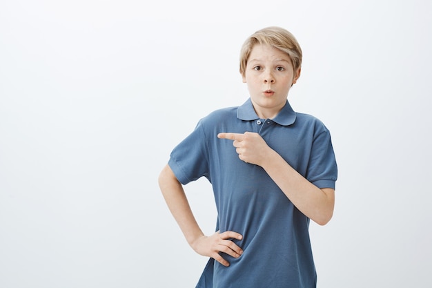 Tir Intérieur De Charmant Garçon Blond étonné Nerveux En T-shirt Bleu, Tenant La Main Sur La Hanche Et Pointant Vers La Gauche Avec L'index