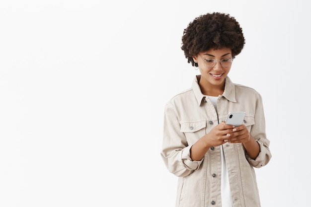 Tir intérieur de charmante jeune femme calme et heureuse dans des verres avec une coiffure afro en tapant un message dans un smartphone regardant avec un sourire joyeux à l'écran du smartphone en lisant un article intéressant sur le web