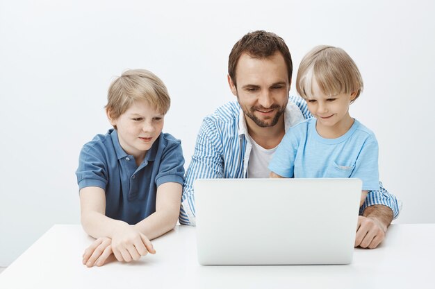 Tir intérieur d'un enfant blond heureux positif avec le père et le frère assis à table, regardant l'écran de l'ordinateur portable et souriant largement