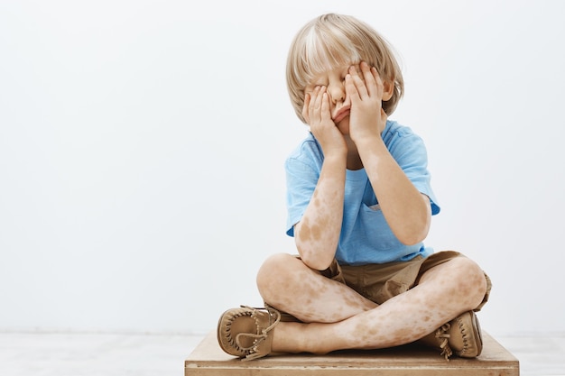 Photo gratuite tir intérieur d'un enfant européen mignon avec une belle coupe de cheveux et vitiligo, couvrant le visage avec des paumes en position assise, jouant à cache-cache avec son frère aîné