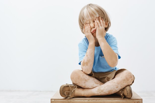 Tir intérieur d'un enfant européen mignon avec une belle coupe de cheveux et vitiligo, couvrant le visage avec des paumes en position assise, jouant à cache-cache avec son frère aîné