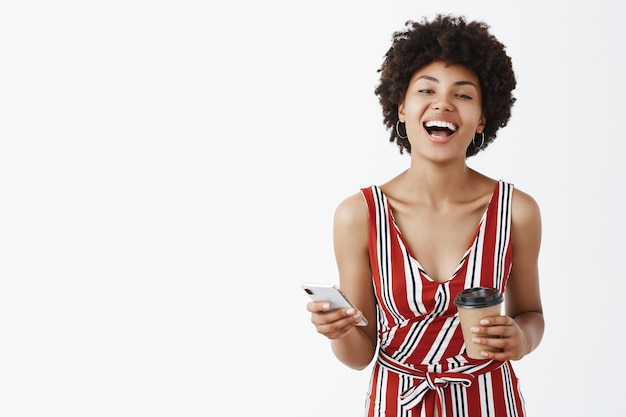 Tir Intérieur D'une Femme Africaine Joyeuse Et émotive Avec Une Coiffure Afro Riant Joyeusement à Haute Voix De Blague Drôle Tenant Une Tasse De Papier Et Un Smartphone S'amusant