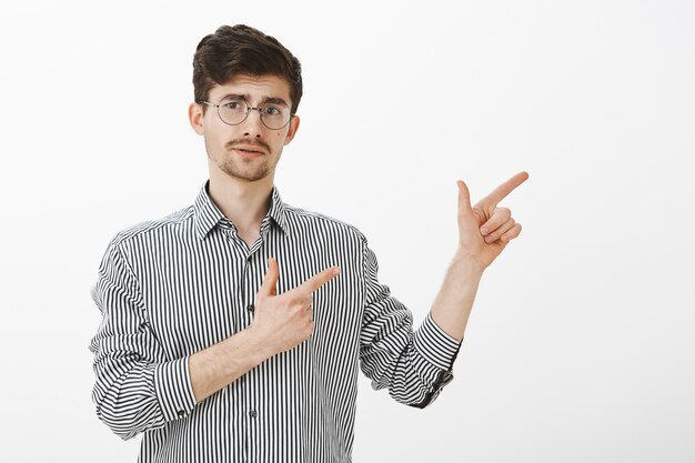 Tir à l'intérieur d'un homme barbu caucasien adulte insécurisé avec moustache dans des lunettes rondes à la mode, pointant vers la droite avec un geste de pistolet, levant les sourcils curieusement, incertain tout en montrant la direction