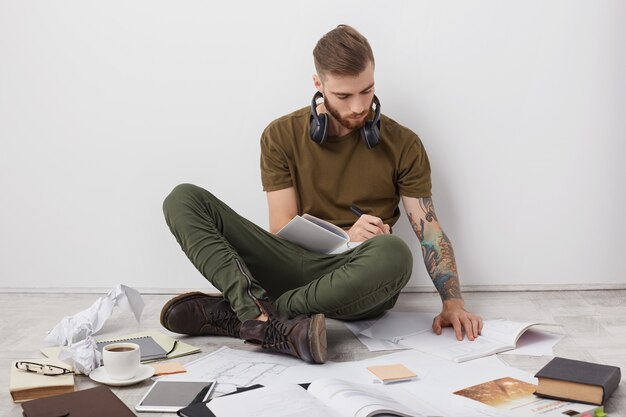 Tir à l'intérieur d'un homme hipster avec une coiffure à la mode, une bérade épaisse et des bras tatoués, regarde attentivement le livre