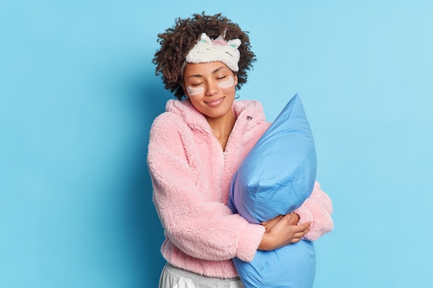 Tir à l'intérieur d'une jeune femme afro-américaine positive pose avec les yeux fermés sourires embrasse doucement oreiller doux