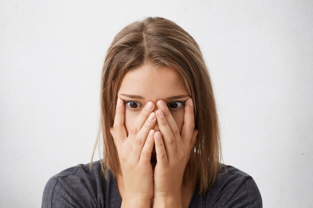 Tir intérieur d'une jeune femme choquée, effrayée ou frustrée couvrant le visage avec les mains, les yeux pleins de terreur et de panique