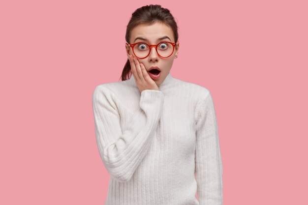 Tir intérieur d'une jeune femme européenne effrayée garde la mâchoire baissée, touche les joues, porte un pull blanc décontracté