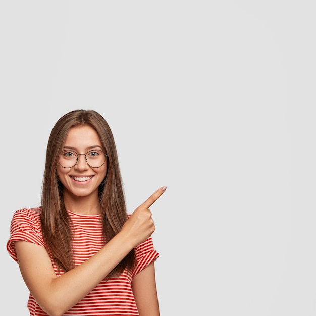 Photo gratuite tir intérieur d'une jeune femme mignonne européenne aux cheveux raides foncés, charmant sourire, pointe de côté avec l'index