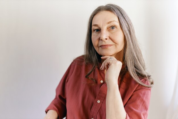 Tir isolé de belle femme senior caucasienne réfléchie avec de longs cheveux gris posant au mur blanc avec un espace de copie pour votre contenu, tenant la main sous le menton, ayant une expression faciale pensif