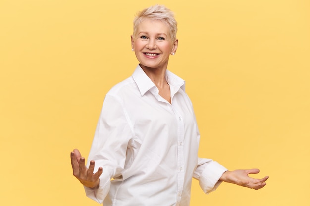 Tir isolé du patron de femme mûre prospère et heureux en chemise blanche tenant les mains écartées et souriant joyeusement, donnant un discours de motivation, dynamisant les employés, sa posture exprimant la confiance