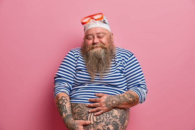 Tir isolé d'un homme barbu dodu tient son gros ventre tatoué ferme les yeux avec satisfaction vêtu d'une chemise de marin sous-dimensionnée, un chapeau de bain en caoutchouc et des lunettes aime la plongée en apnée pendant l'été