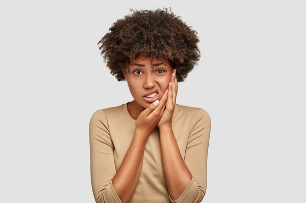 Tir isolé d'une jeune femme à la peau sombre mécontente a une coupe de cheveux afro, garde la main sur la joue, fronce les sourcils de douleur, souffre de maux de dents, vêtue d'un pull décontracté, exprime des émotions négatives