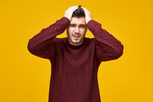 Tir isolé d'un jeune homme frustré en pull tricoté gardant les mains sur la tête et grimaçant souffrant de migraine en raison d'un travail stressant, ayant une expression faciale douloureuse, se sentant mal