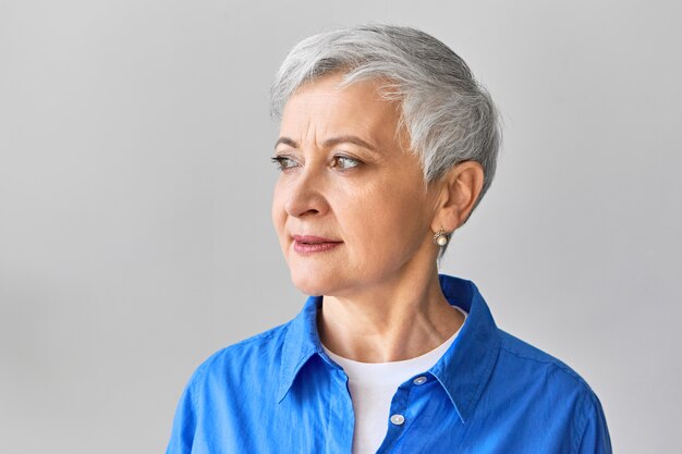 Tir isolé de la jolie femme aux cheveux gris de soixante ans portant des boucles d'oreilles en perles et une chemise bleue sur un haut blanc en détournant les yeux avec une expression faciale sérieuse songeuse. Concept de personnes et de style de vie