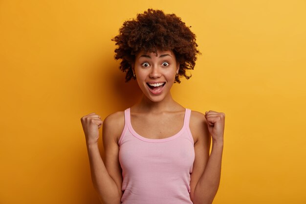 Tir isolé d'une joyeuse femme afro-américaine à la peau sombre serre les poings avec triomphe, heureuse de devenir champion et de remporter le premier prix, sourit joyeusement, applaudit les nouvelles positives, porte un gilet décontracté