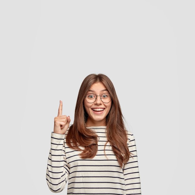 Tir isolé de joyeuse femme de race blanche aux cheveux raides, montre l'espace libre, points avec l'index pour votre contenu publicitaire, porte un pull rayé décontracté, isolé sur un mur blanc