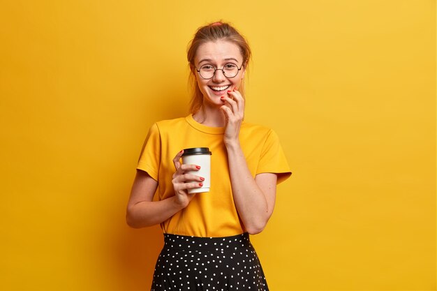 Tir isolé de joyeuse fille sourit exprime joyeusement des sentiments sincères boit du café à emporter