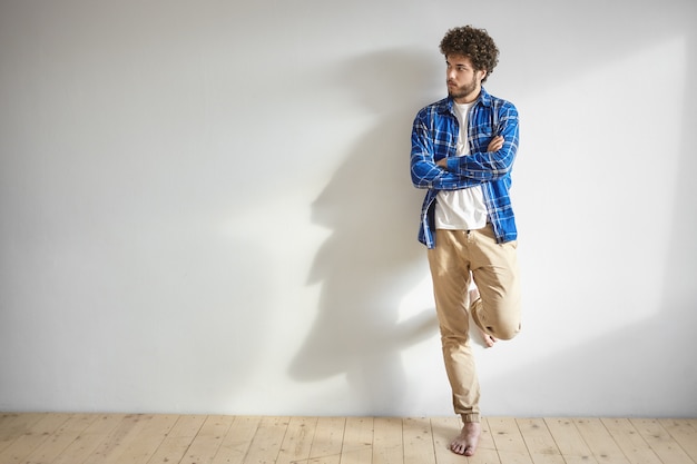 Tir isolé de séduisant jeune homme de race blanche avec une barbe épaisse et les pieds nus posant à l'intérieur debout contre un mur blanc blanc avec copie espace pour votre contenu promotionnel