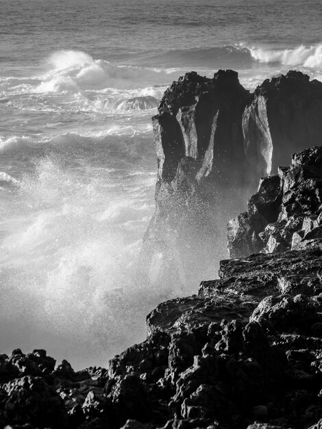 Tir en niveaux de gris de fortes vagues frappant de gros rochers sur une rive avec des éclaboussures d'eau et de pulvérisation