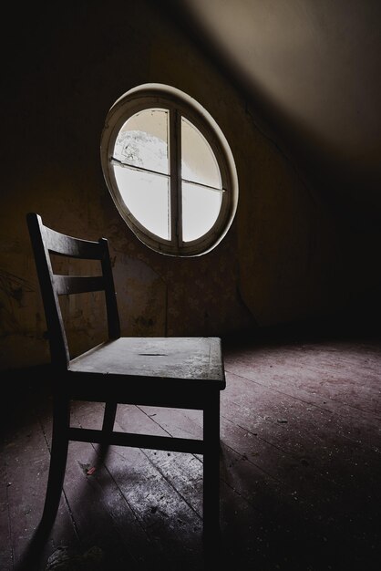 Tir vertical d'une chaise en bois dans une pièce sombre avec une fenêtre ronde - concept d'isolement