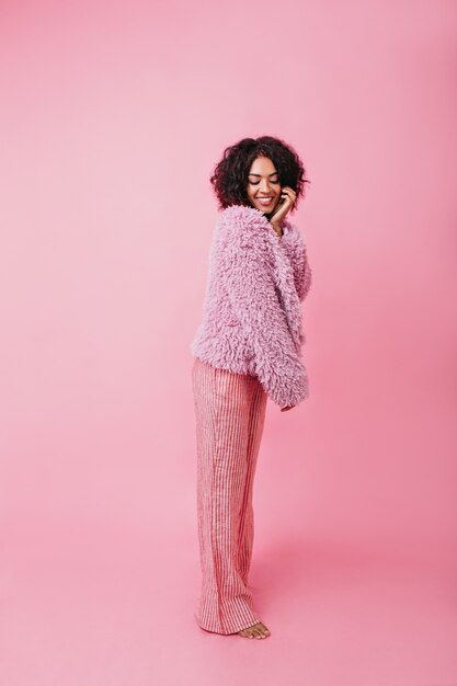 Tir vertical confortable de couleur rose. Fille aux pieds nus en pantalon large rayé et manteau de fourrure touche son visage avec raideur.