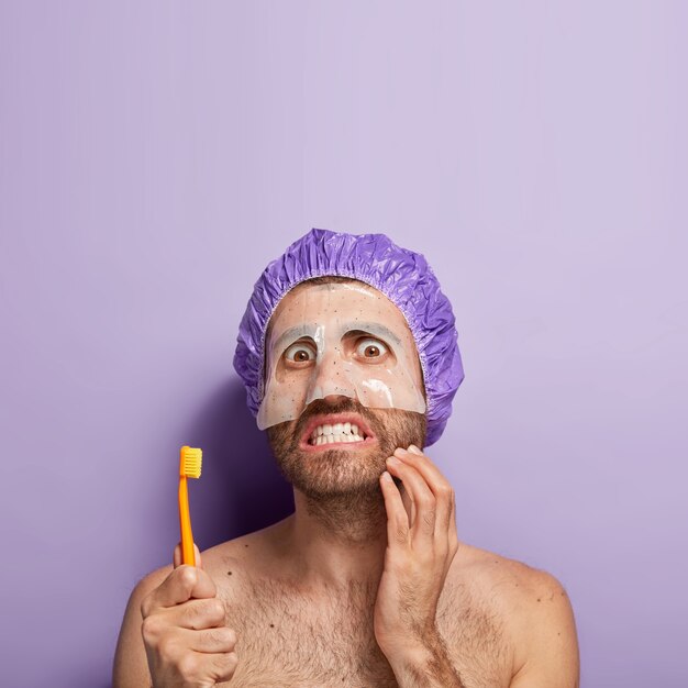 Tir vertical du beau modèle masculin touche des poils épais, porte un masque hydratant pour la douceur de la peau, serre les dents, tient la brosse à dents